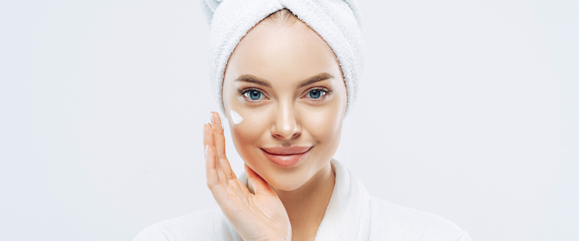 A woman with a towel wrapped on her head is smiling while applying face cream to her cheek.