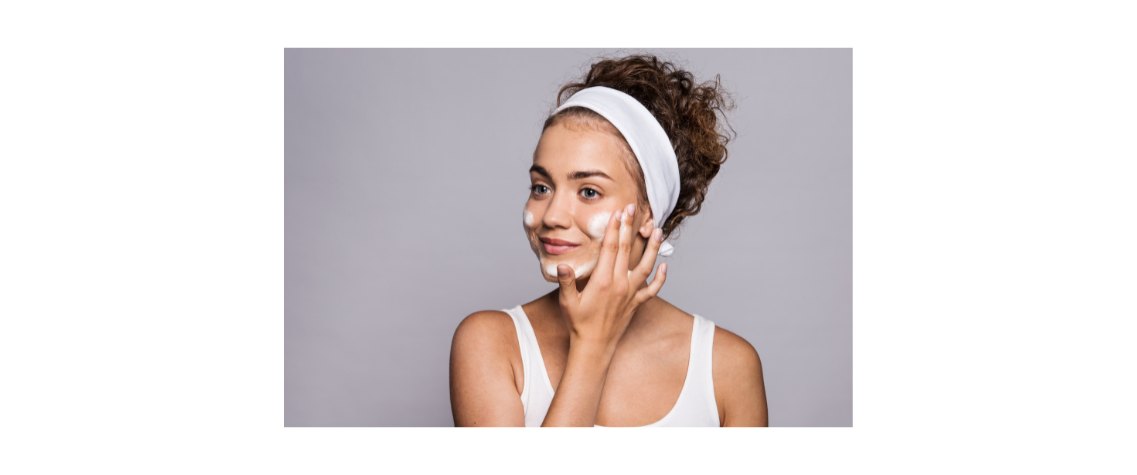 A young woman with curly hair and a headband is smiling while applying cream to her cheeks as part of her skincare routine.A young woman with curly hair and a headband is smiling while applying cream to her cheeks as part of her skincare routine.